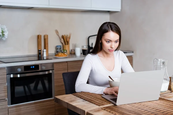 Attraente freelance utilizzando laptop vicino brocca con latte a casa — Foto stock