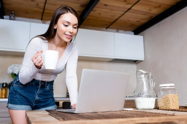 Selective focus of happy freelancer using laptop and holding cup with drink — Stock Photo