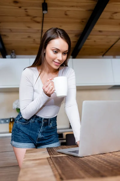 Selective focus of happy freelancer using laptop and holding cup with tea — Stock Photo