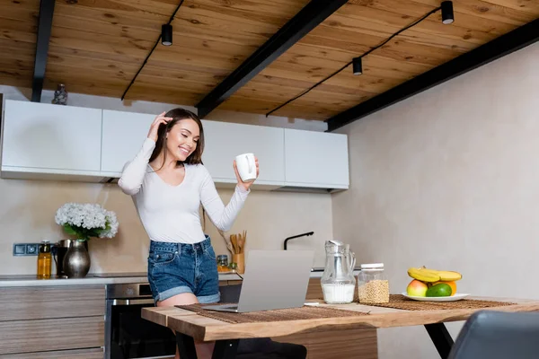 Feliz freelancer sosteniendo taza cerca del ordenador portátil, jarra con leche y frutas en la cocina - foto de stock