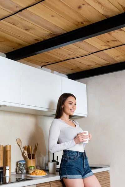 Heureuse fille tenant tasse de thé près de la plaque avec croissant sur la table — Photo de stock