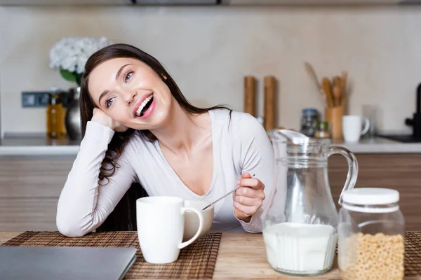 Fille gaie tenant cuillère près du bol, tasse et cruche avec du lait — Photo de stock