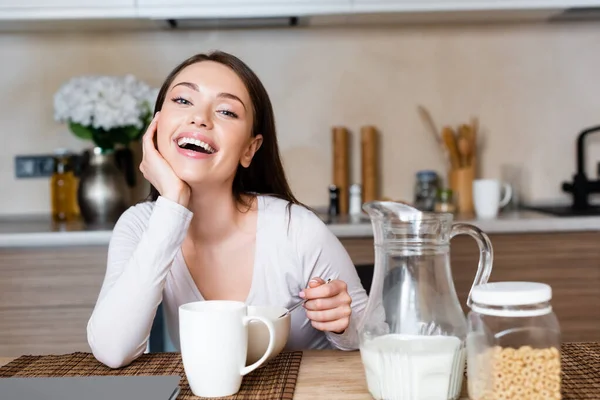 Счастливая девушка держит ложку возле миски, чашку и кувшин с молоком — стоковое фото