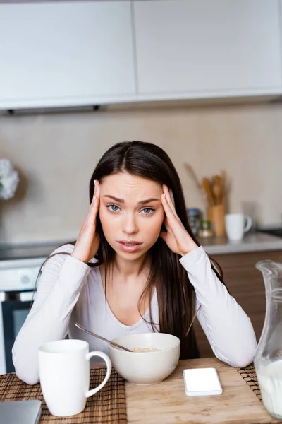 Upset girl touching head near bowl, jug and smartphone with white screen — Stock Photo