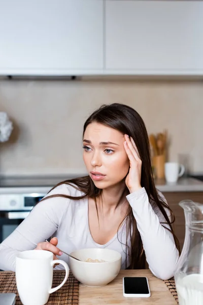 Sad girl touching head near bowl, jug and smartphone with blank screen — Stock Photo