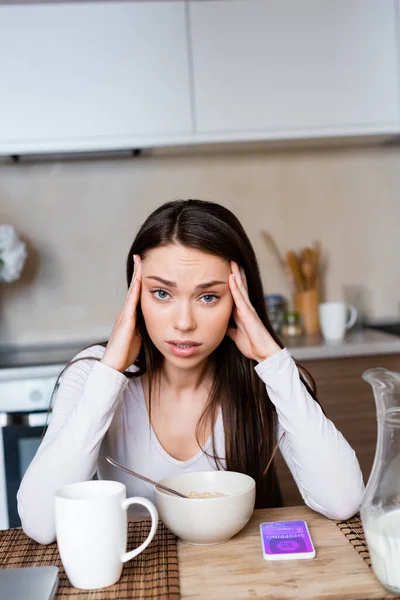 Upset girl touching head near bowl, jug and smartphone with online shopping app — Stock Photo
