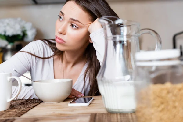 Selective focus of sad girl near bowl, jug, cup and smartphone with blank screen — Stock Photo