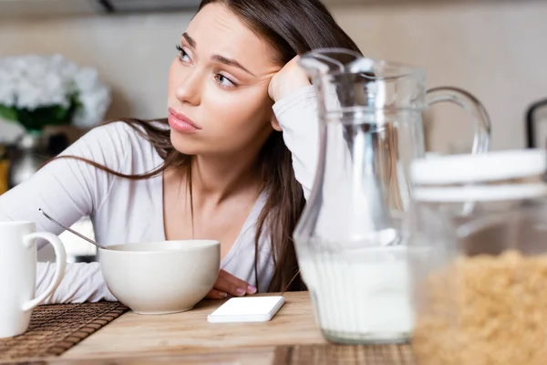 Selective focus of sad girl near bowl, jug, cup and smartphone with white screen — Stock Photo