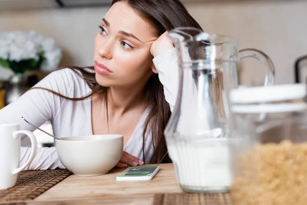 Selective focus of upset girl near bowl, jug, cup and smartphone with booking app — Stock Photo