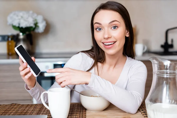 Enfoque selectivo de la chica feliz apuntando con el dedo al teléfono inteligente con pantalla en blanco cerca de la taza, tazón y jarra con leche - foto de stock
