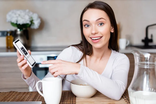 KYIV, UCRAINA - 29 APRILE 2020: focus selettivo della ragazza felice che punta il dito contro lo smartphone con app uber vicino a tazza, ciotola e brocca con latte — Foto stock