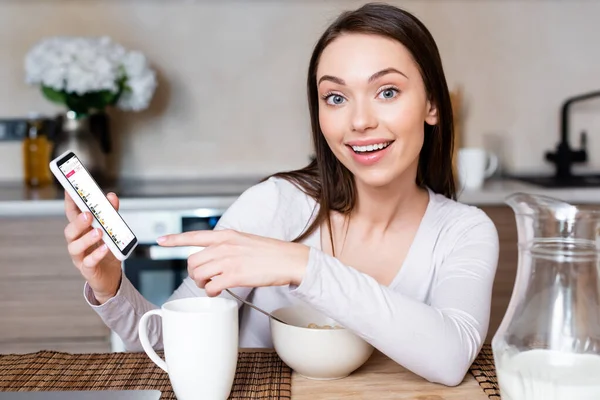 KYIV, UCRANIA - 29 DE ABRIL DE 2020: enfoque selectivo de la chica feliz apuntando con el dedo al teléfono inteligente con la aplicación de música de manzana cerca de la taza, tazón y jarra con leche - foto de stock