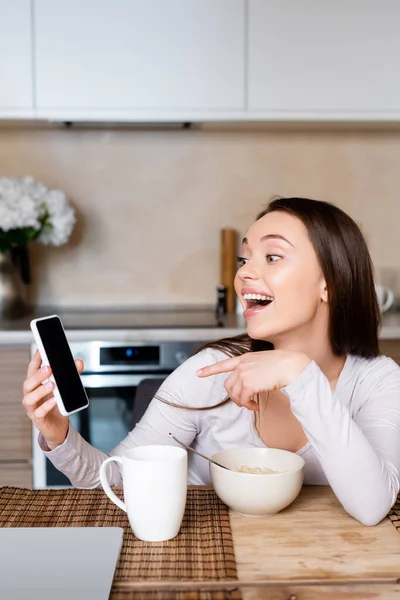 Chica emocionada señalando con el dedo en el teléfono inteligente con pantalla en blanco cerca de taza y tazón - foto de stock