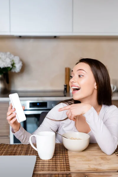 Chica emocionada apuntando con el dedo en el teléfono inteligente con pantalla blanca cerca de taza y tazón - foto de stock