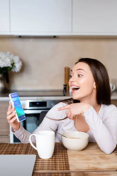 Mujer emocionada señalando con el dedo en el teléfono inteligente con aplicación de reserva cerca de taza y tazón - foto de stock