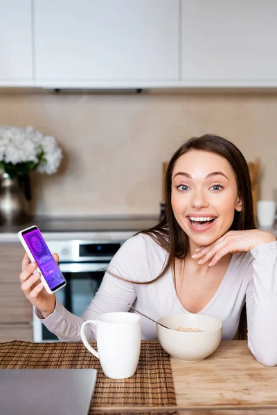 Збуджена жінка тримає смартфон із додатком для покупок в Інтернеті біля чашки та миски — стокове фото