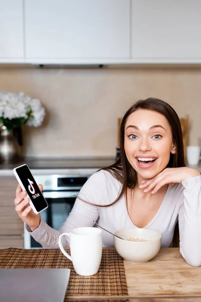 KYIV, UKRAINE - APRIL 29, 2020: excited woman holding smartphone with tik tok app near cup and bowl — Stock Photo