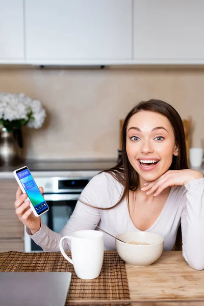 Femme excitée tenant smartphone avec application de réservation près de tasse et bol — Photo de stock