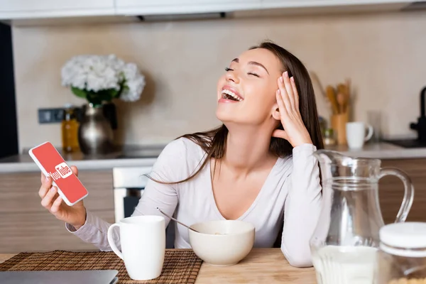 KYIV, UKRAINE - APRIL 29, 2020: happy woman holding smartphone with youtube app and laughing near breakfast — Stock Photo