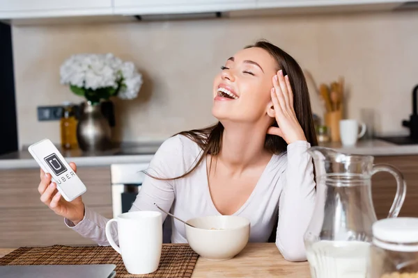 KYIV, UKRAINE - 29 de abril de 2020: mulher feliz segurando smartphone com aplicativo uber e rindo perto do café da manhã — Fotografia de Stock