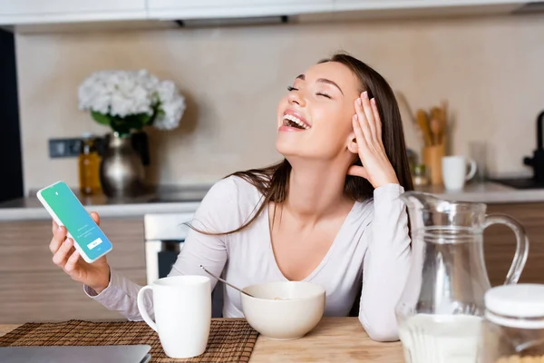 KYIV, UCRANIA - 29 de abril de 2020: mujer feliz sosteniendo el teléfono inteligente con la aplicación de twitter y riendo cerca del desayuno - foto de stock