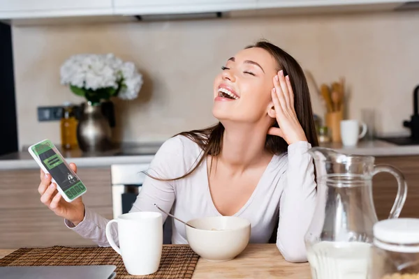 Mujer feliz sosteniendo teléfono inteligente con aplicación de reserva y riendo cerca del desayuno - foto de stock