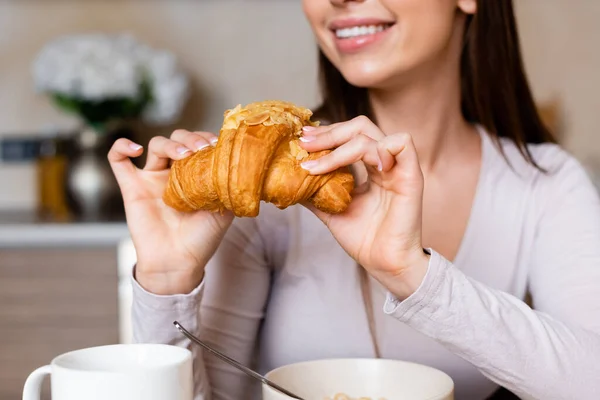 Vista recortada de chica feliz sosteniendo croissant sabroso cerca de cuenco y taza - foto de stock
