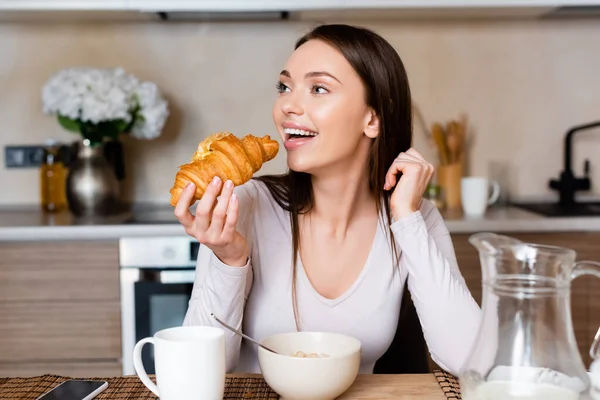 Enfoque selectivo de chica alegre sosteniendo croissant cerca de teléfono inteligente con pantalla en blanco - foto de stock