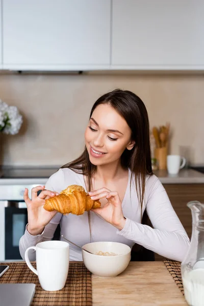 Fröhliches Mädchen blickt auf leckeres Croissant in der Nähe von Schüssel und Tasse — Stockfoto