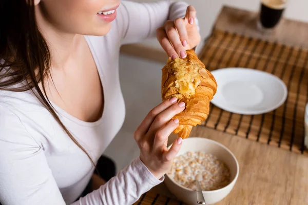 Vista recortada de chica alegre sosteniendo croissant cerca de tazón con hojuelas de maíz - foto de stock
