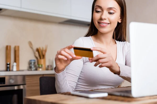 Selective focus of cheerful girl holding credit card near laptop and smartphone on table — Stock Photo