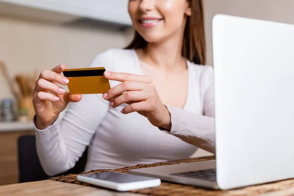 Vista cortada de menina feliz segurando cartão de crédito perto de laptop e smartphone na mesa — Fotografia de Stock