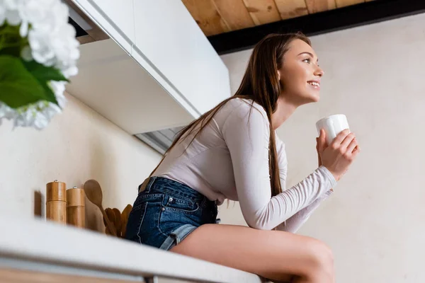 Messa a fuoco selettiva di sorridente ragazza seduta e in possesso di tazza di caffè — Foto stock