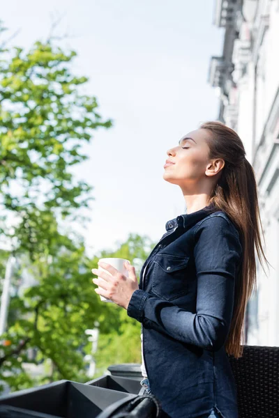 Vue latérale de la jeune femme joyeuse avec les yeux fermés tenant tasse de café à l'extérieur — Photo de stock