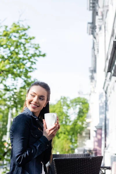 Sonriente mujer joven en chaqueta de mezclilla sosteniendo la taza de café fuera - foto de stock