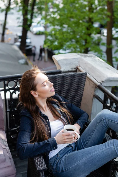 Menina alegre em jeans jeans jeans e casaco sentado na cadeira e segurando xícara de café na varanda — Fotografia de Stock