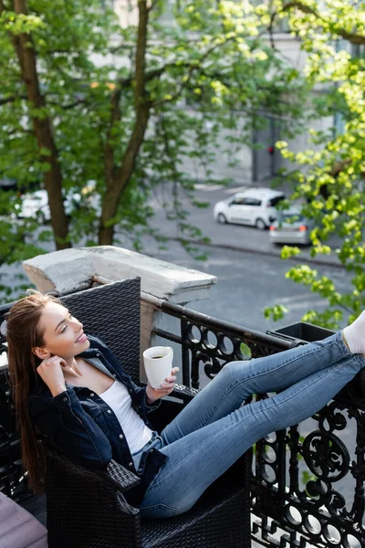 Chica sonriente en vaqueros y chaqueta sentado en la silla y la celebración de la taza de café en el balcón - foto de stock