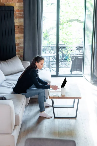 Atractivo freelancer con ordenador portátil con pantalla en blanco cerca de la taza de café en la mesa de café - foto de stock