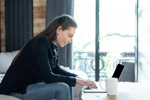 Side view of happy freelancer using laptop with blank screen near cup on coffee table — Stock Photo