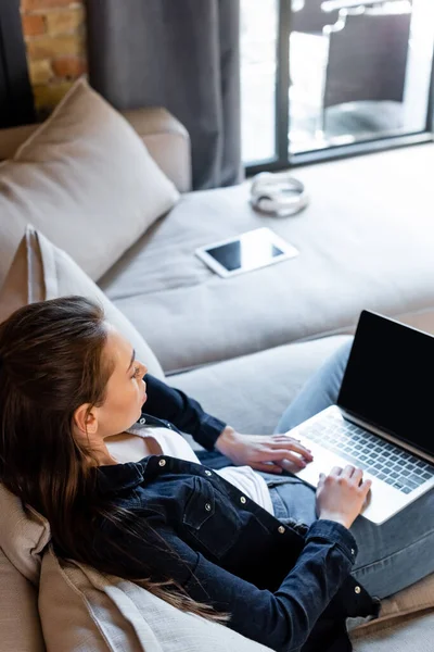 Enfoque selectivo de joven freelancer usando portátil con pantalla en blanco cerca de la tableta digital en el sofá - foto de stock