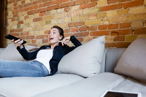 Surprised girl holding remote controller and sitting on sofa — Stock Photo