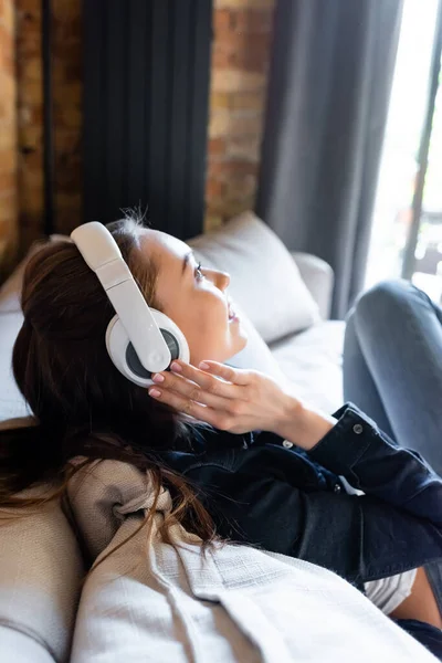 Vista lateral de chica alegre tocando auriculares inalámbricos mientras escucha música - foto de stock