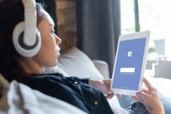 KYIV, UCRAINA - 29 APRILE 2020: focus selettivo della ragazza in cuffie wireless ascoltare musica e utilizzando tablet digitale con applicazione facebook — Foto stock