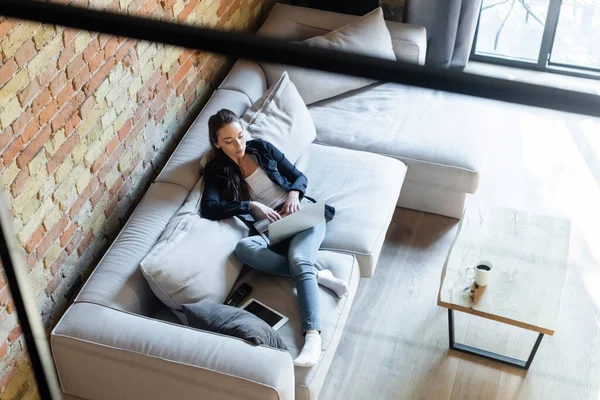 Selective focus of attractive freelancer using laptop near digital tablet with blank screen on sofa — Stock Photo