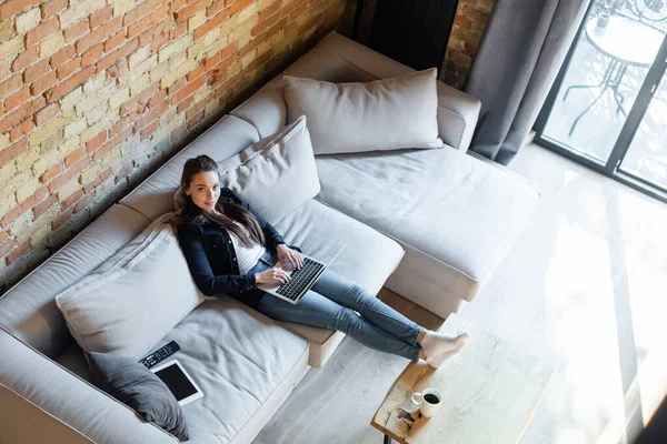 Vista aérea del atractivo freelancer utilizando un ordenador portátil cerca de la tableta digital con pantalla en blanco en el sofá - foto de stock