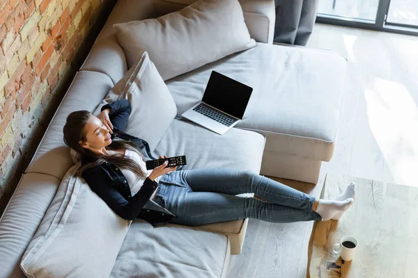 Vue aérienne de la femme heureuse tenant télécommande près de l'ordinateur portable avec écran blanc sur le canapé et la tasse sur la table basse — Photo de stock