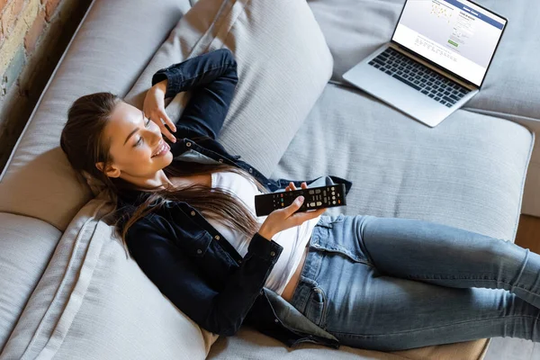 KYIV, UKRAINE - APRIL 29, 2020: overhead view of happful woman holding remote controller near laptop with facebook website on sofa — стокове фото