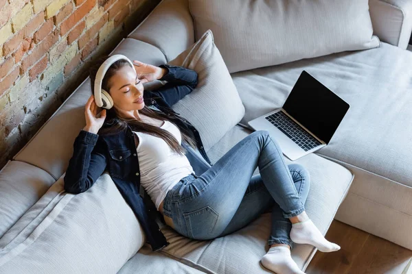 Vista aerea di giovane donna con gli occhi chiusi ascoltare musica in cuffie wireless vicino al computer portatile con schermo bianco — Foto stock