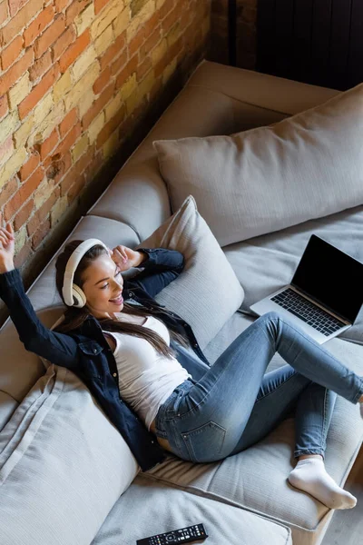 Vista aérea de la mujer feliz escuchando música en auriculares inalámbricos cerca de la computadora portátil con pantalla en blanco - foto de stock