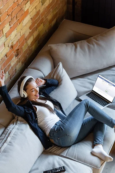 KYIV, UCRANIA - 29 de abril de 2020: vista aérea de una mujer feliz escuchando música en auriculares inalámbricos cerca de la computadora portátil con el sitio web de facebook - foto de stock